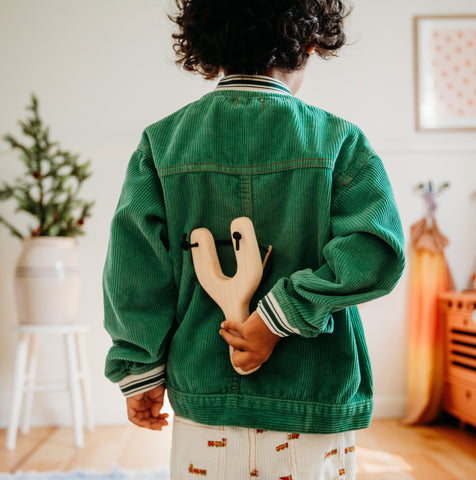 Child with dark, curly hair in green corduroy jacket holds natural colored wooden slingshot in his hand behind his back. 