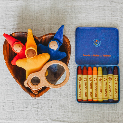 A heart shaped wooden bowl with three small star baby pocket waldorf dolls in red, gold, and blue and a multiple lens magnifying glass. Next to the bowl is a tin of 8 stockmar beeswax stick crayons in waldorf color assortment