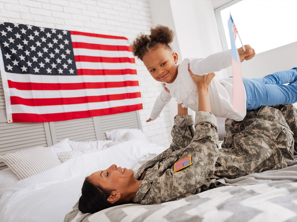 Woman in military uniform laying on a bed holding her daughter up and smiling