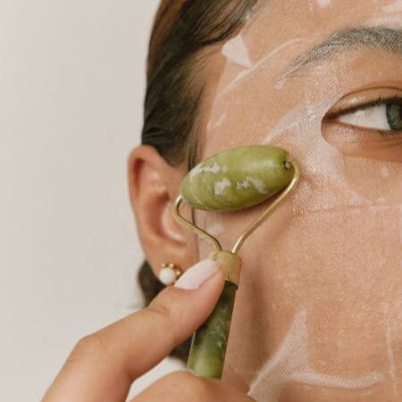 close up woman face jade roller depuff over sheet mask