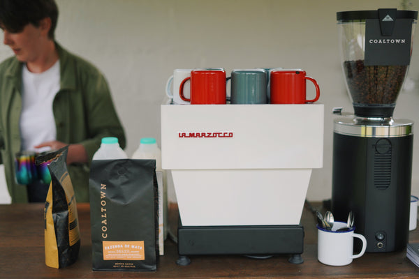 Making coffee with a La Marzocco Linea Mini. Pictured is a coffee grinder, enamel mugs, bottles of milk and a bag of Coaltown Fazenda de Mata coffee. 