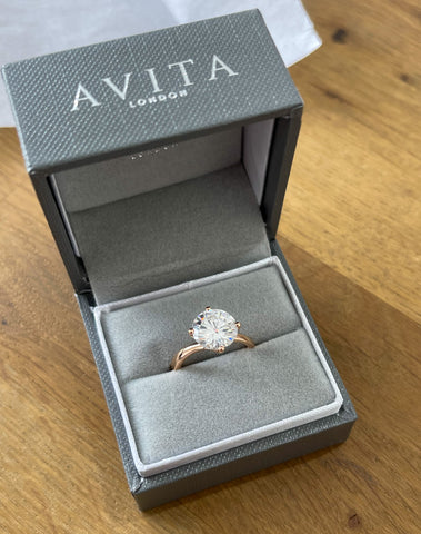 An Avita Jewellery grey ring box containing a large round diamond engagement ring with an elegant gold twist band.