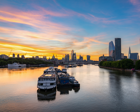 a blue, pink and golden sunset over the river thames with London's skyline in the distance and two medium-large boats floating on the water in the foreground