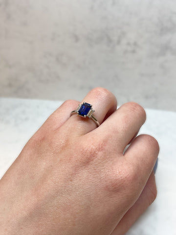 A women's hand wearing a blue sapphire trilogy engagement ring. Rich blue, rectangular, emerald-cut sapphire centre stone with a baguette-cut rectangular diamond on either side