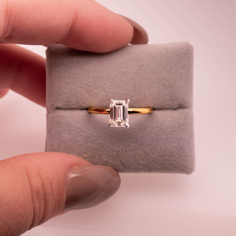 Emerald-cut diamond gold solitaire engagement ring being held within a grey ring cushion.