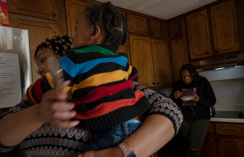 ic:Angelica Lyons carries her son while her younger sister, Ansonia Lyons, finishes her snack in the background.