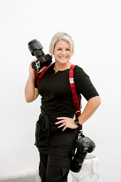 Photographer holding a camera and wearing a holdfast strap with another camera and wearing a black linen apron.