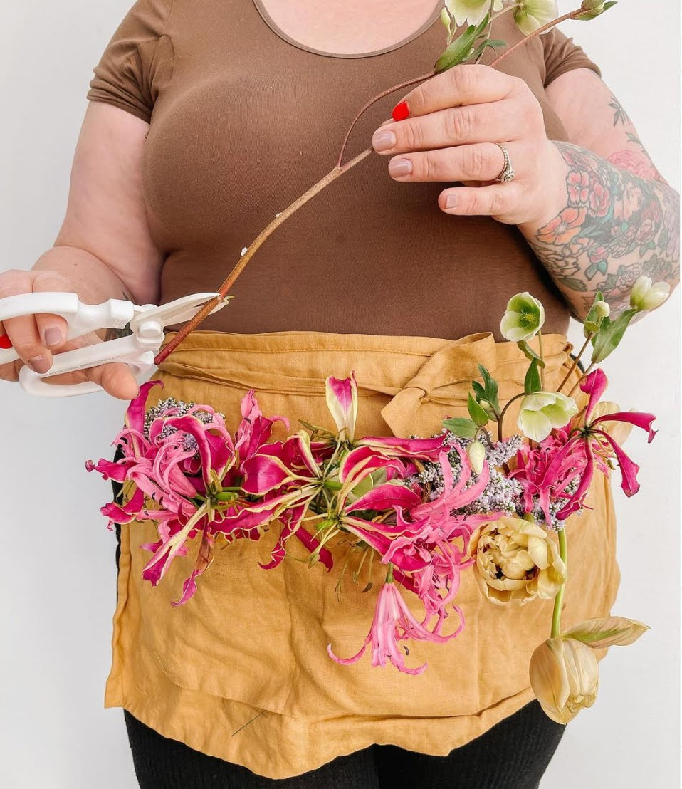 Becky clipping the ends of a stem and wearing Gather 33 linen apron in gold that is filled with pink and green flowers.