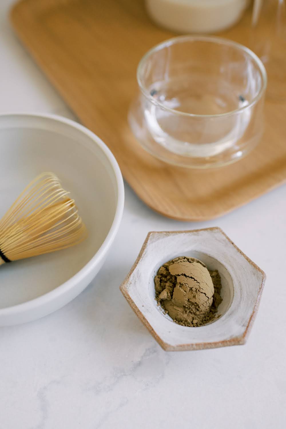 Hojicha powder in a hexagon ceramic bowl beside a ceramic bowl with a Japanese tea whisk and a wooden tray with cup of water.