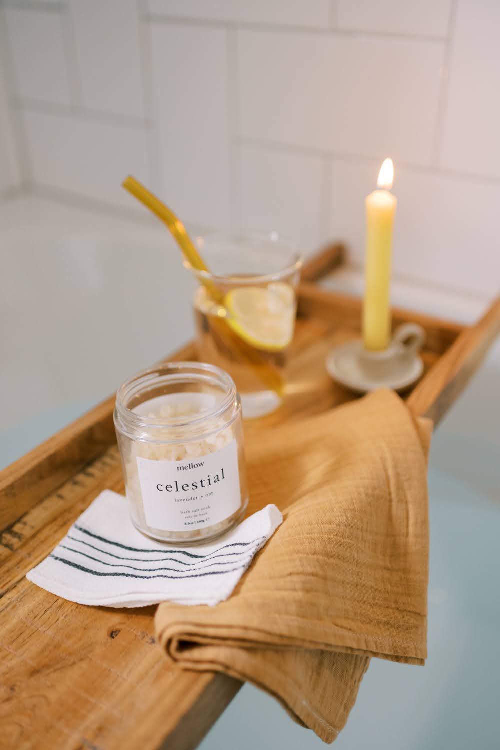 Bath caddy with bath soak, exfoliating mitt, hand towel, glass of water with glass straw and a lit beeswax candle.
