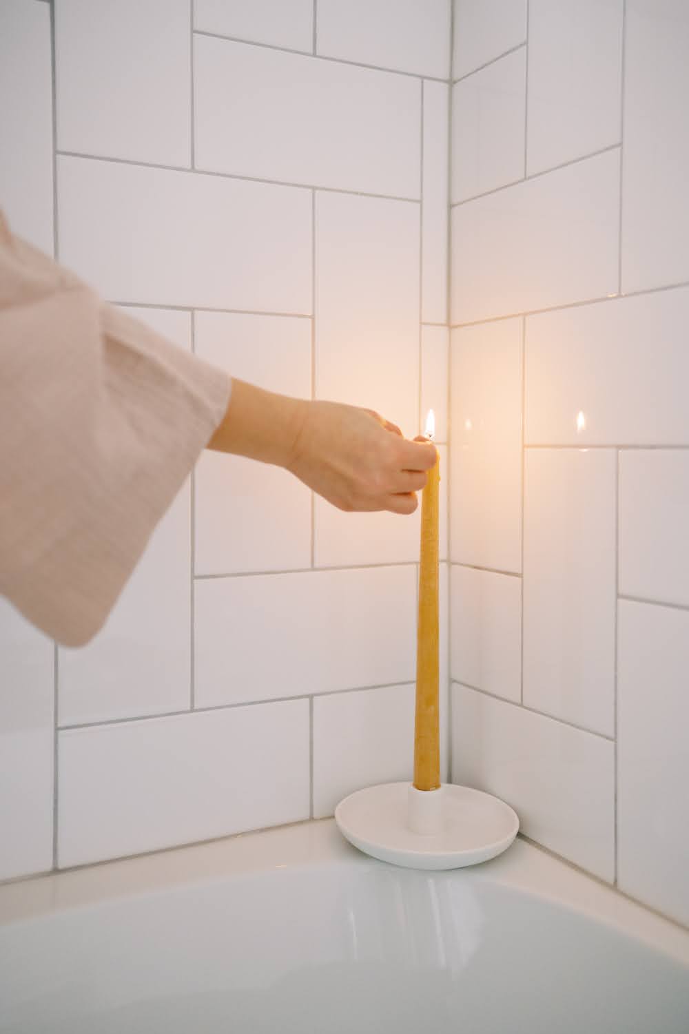 Woman's hand in blush bathrobe lighting a tapered beeswax candle in the bathtub