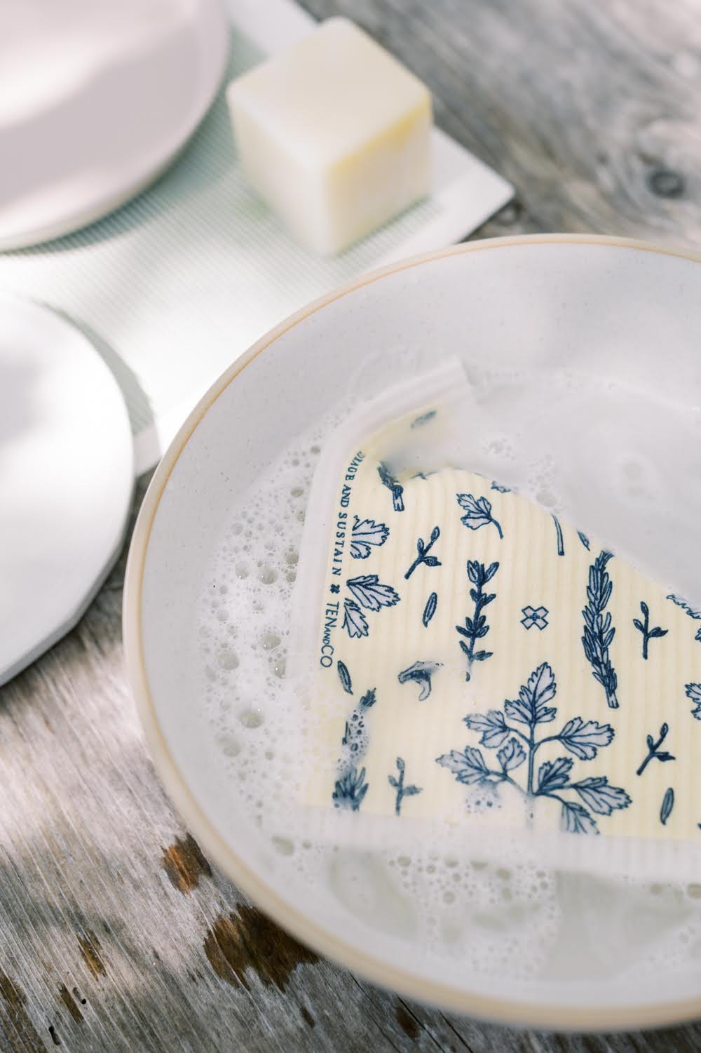 Spongecloth in a bowl of soapy water, with solid dish soap and dishes drying on a spongecloth mat