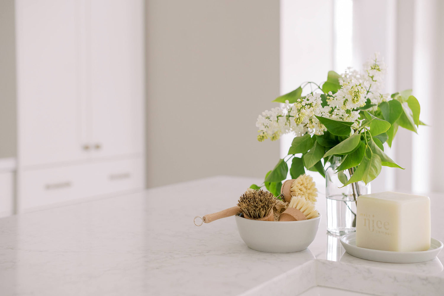 Bright neutral kitchen with sustainable dish brushes in a bowl and a large solid dish soap on the kitchen counter with lilacs in a vase behind them.