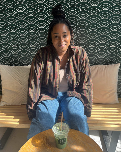 Jordan sitting on a bench at the Matchaful Café drinking an iced matcha with a seigaiha patterned wallpaper behind her