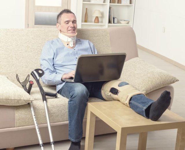 A man sits on the couch working on his laptop with a knee brace on post knee surgery.
