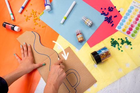 cutting out cardboard sword craft with xacto knife on colorful paper background with glitter, beads, paint, and stickers