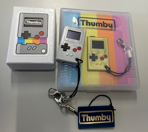 Special edition gold thumby in a case, with a gray Thumby in a standard shipping box with a PCB keychain strewn across the bottom