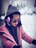 Niño pequeño con snowcaot rosa con fondo de nieve exterior usando gafas de sol para niños banz y protección auditiva para bebés
