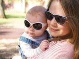 adult womnan holds infant in arms with park background, both wear sunglasses