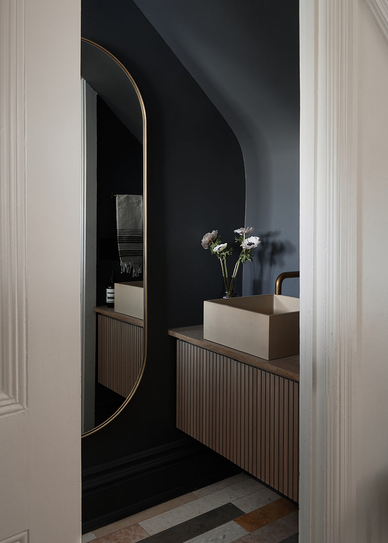 Moody modern bathroom with dark grey walls, a wooden cabinet and sink, and colorful stone tiles on the floor.