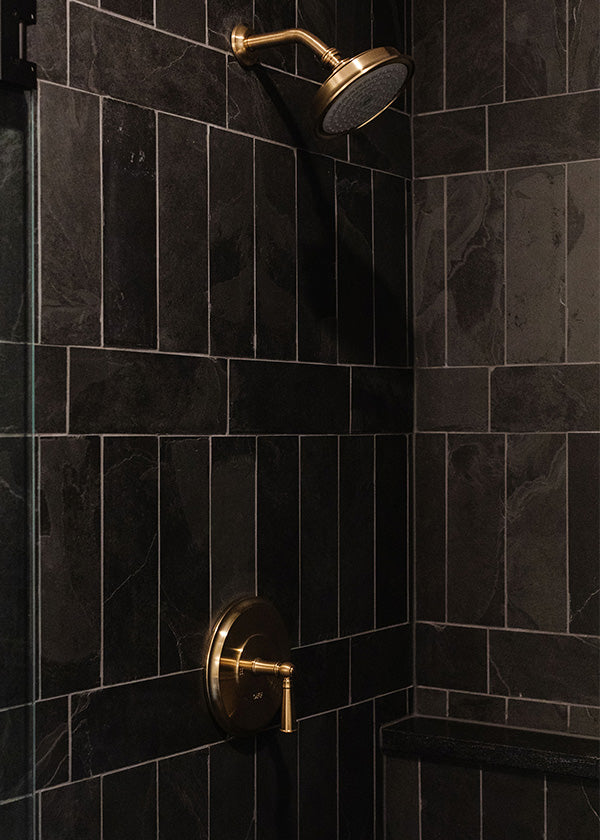 Dark colored shower corner with a slate tiled wall and a brass showerhead and handle.