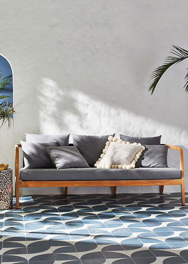 Outdoor living area with white stucco wall, blue and white cement tile floor, and grey wood sofa.