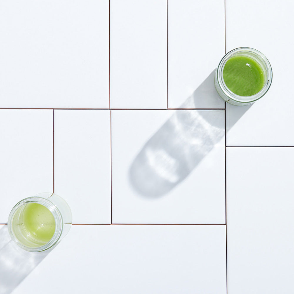 Overhead shot of glossy white ceramic tile with two jars containing green liquid sitting on top.
