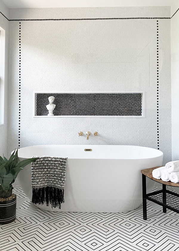 Black and white tiled tub area with penny rounds on the wall, patterned cement tile on the floor, a white tub, and black accents.