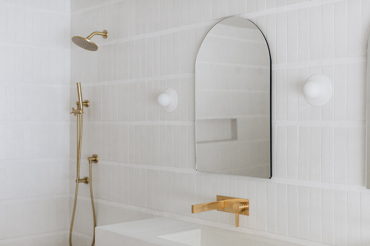 Airy white bathroom vanity area with arched mirrore, glazed brick wall, and brass hardware.