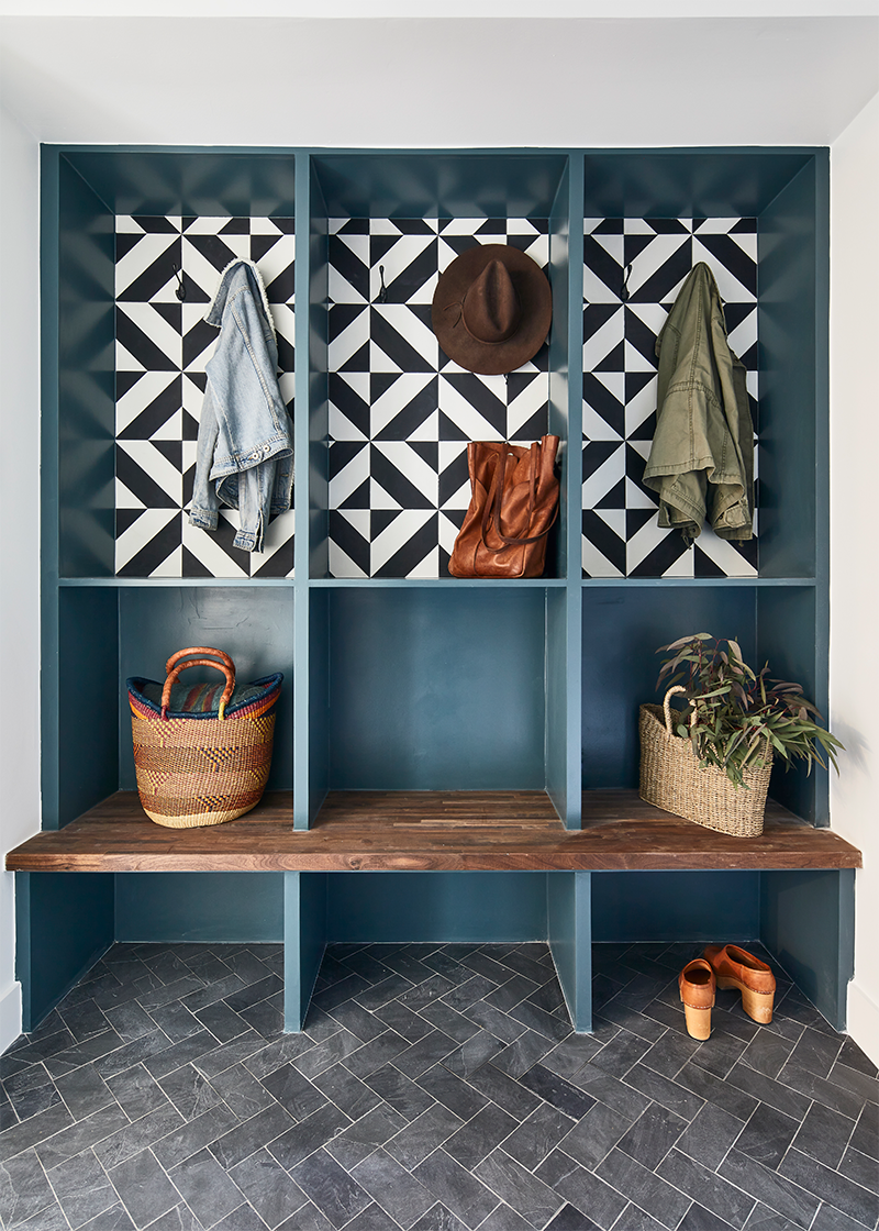 Vignette of mudroom with blue painted walls, a black and white cement tile backsplash, a wooden bench, and a slate tile herringbone floor.