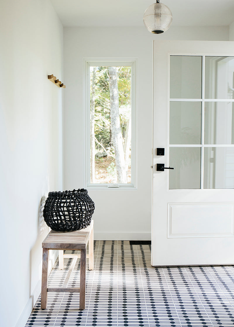 White entryway with wooden bench, white door, and black and white cement patterned tile floor.