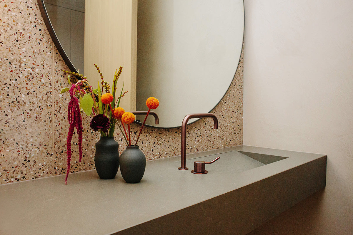 Modern bathroom with tan terrazzo tile backsplash, grey countertop, and large circular mirror.