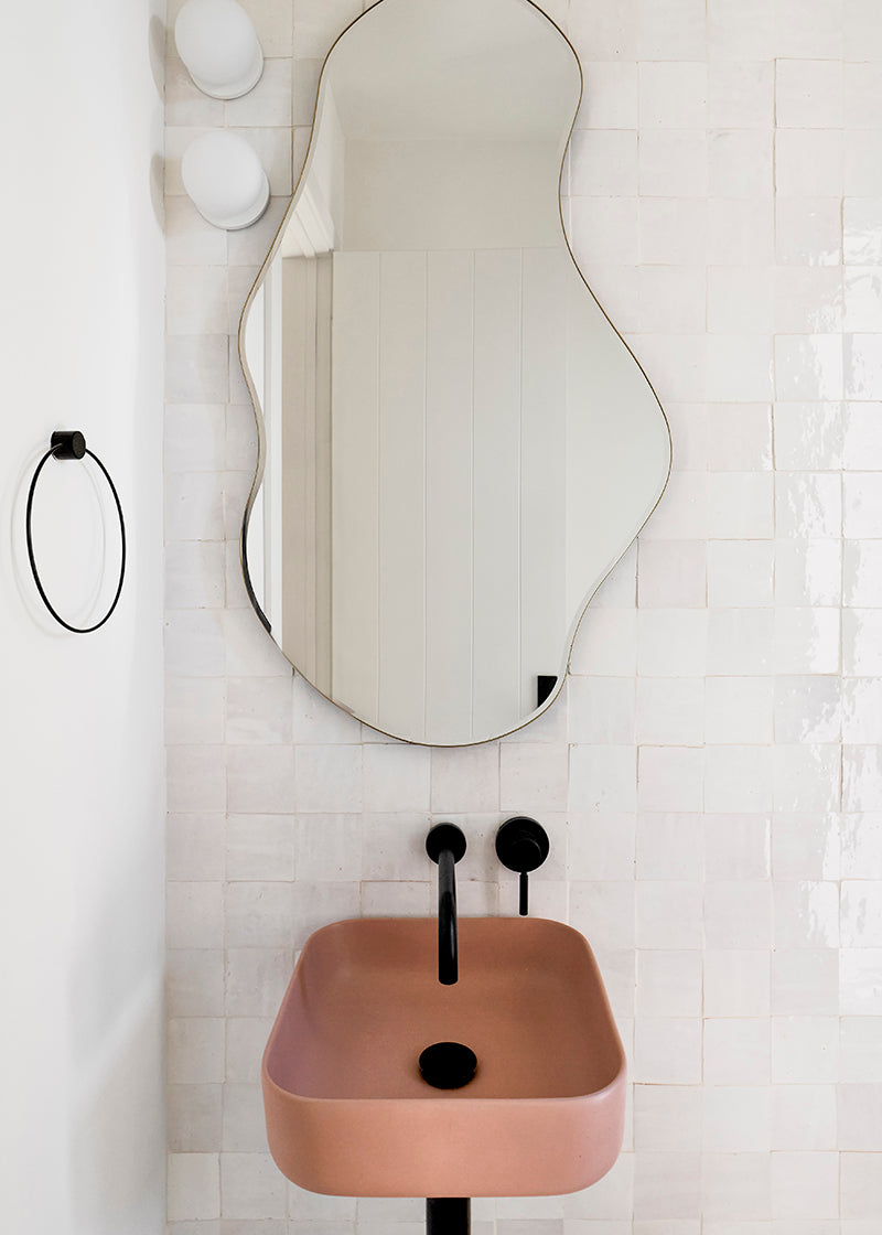 Airy white zellige tile bathroom with curvy mirror and terracotta colored sink.