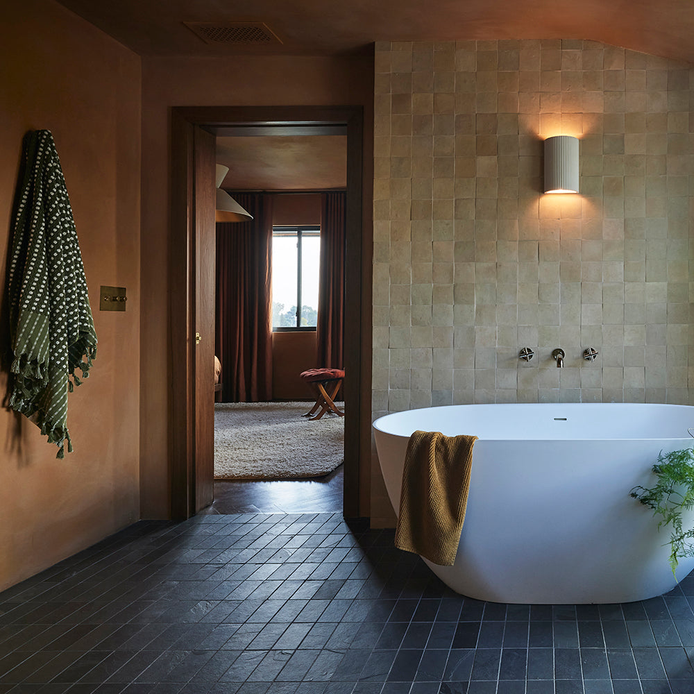 Earth toned bathroom with natural zellige tile wall, slate subway floor, white tub, and green towels.