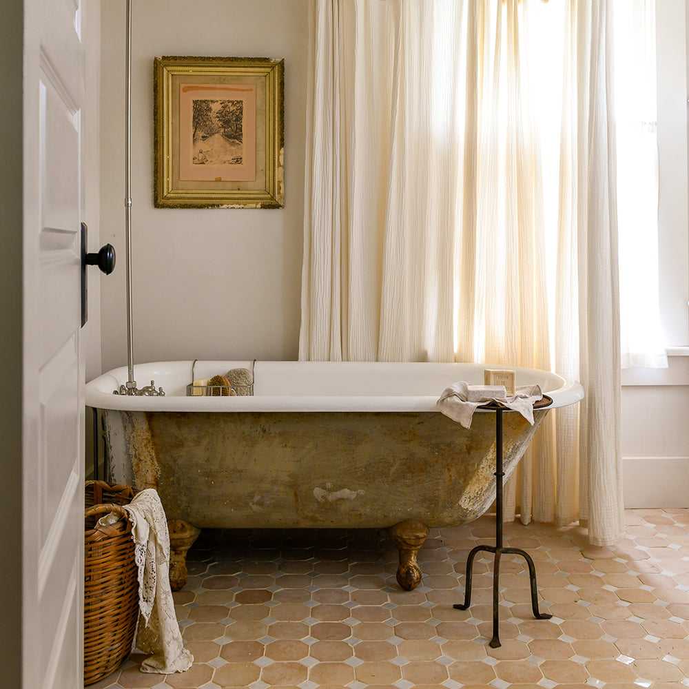 A French inspired bath tub area with an antique claw foot tub, natural terracotta tile floor, and long curtains.