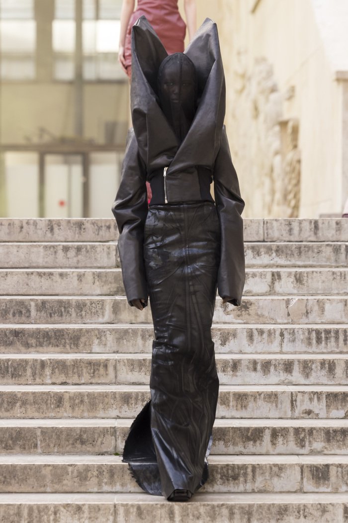 A runway look with a coat with lapels that extend far above the models head on the sides and sleeves that pass her hands.