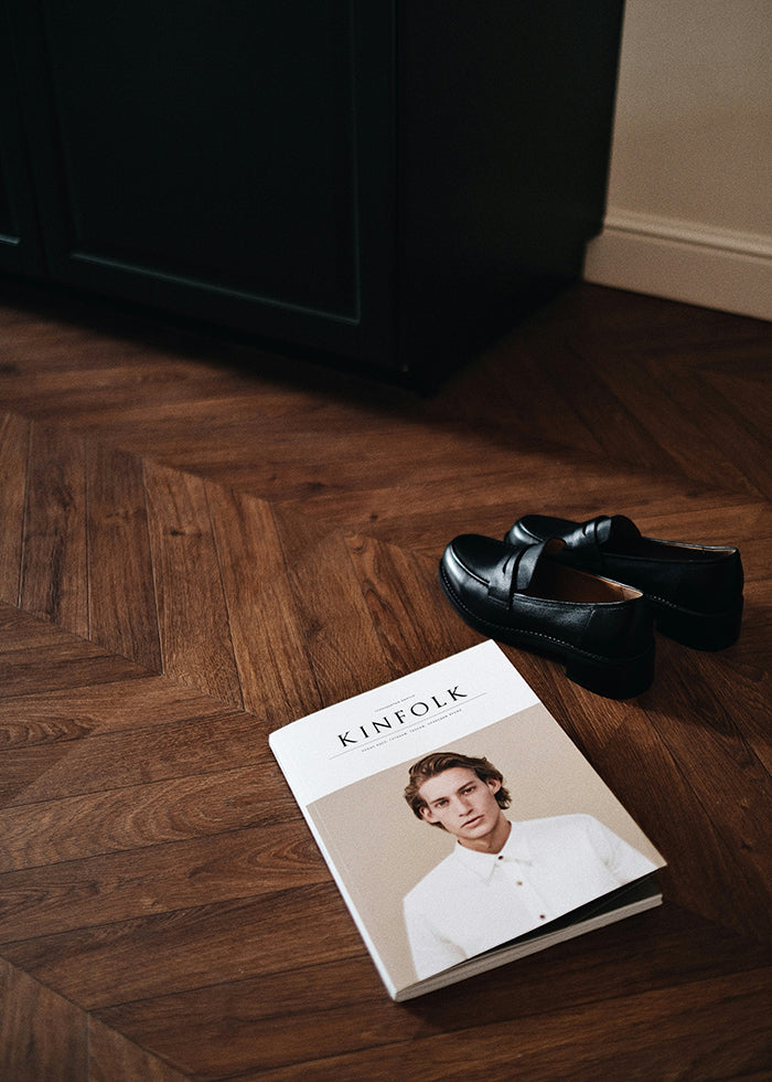 Closeup of a dark wooden chevron floor with black loafers and a magazine.