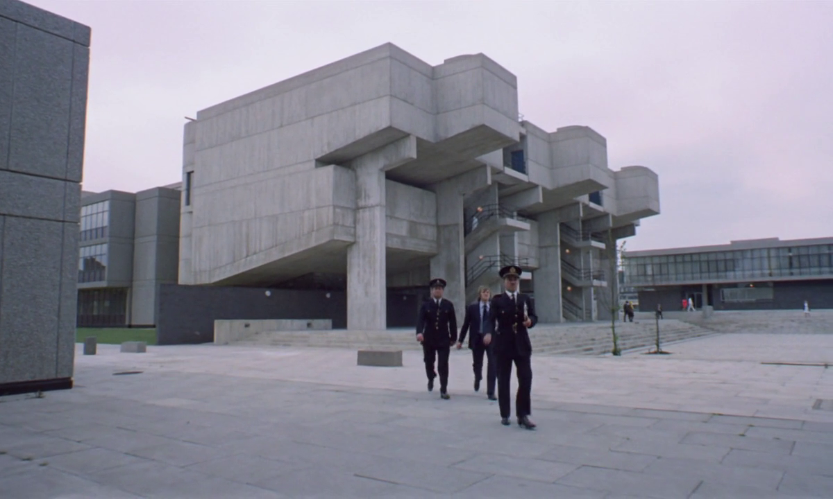 Characters in uniform from A Clockwork Orange walk in front of a brutalist building.