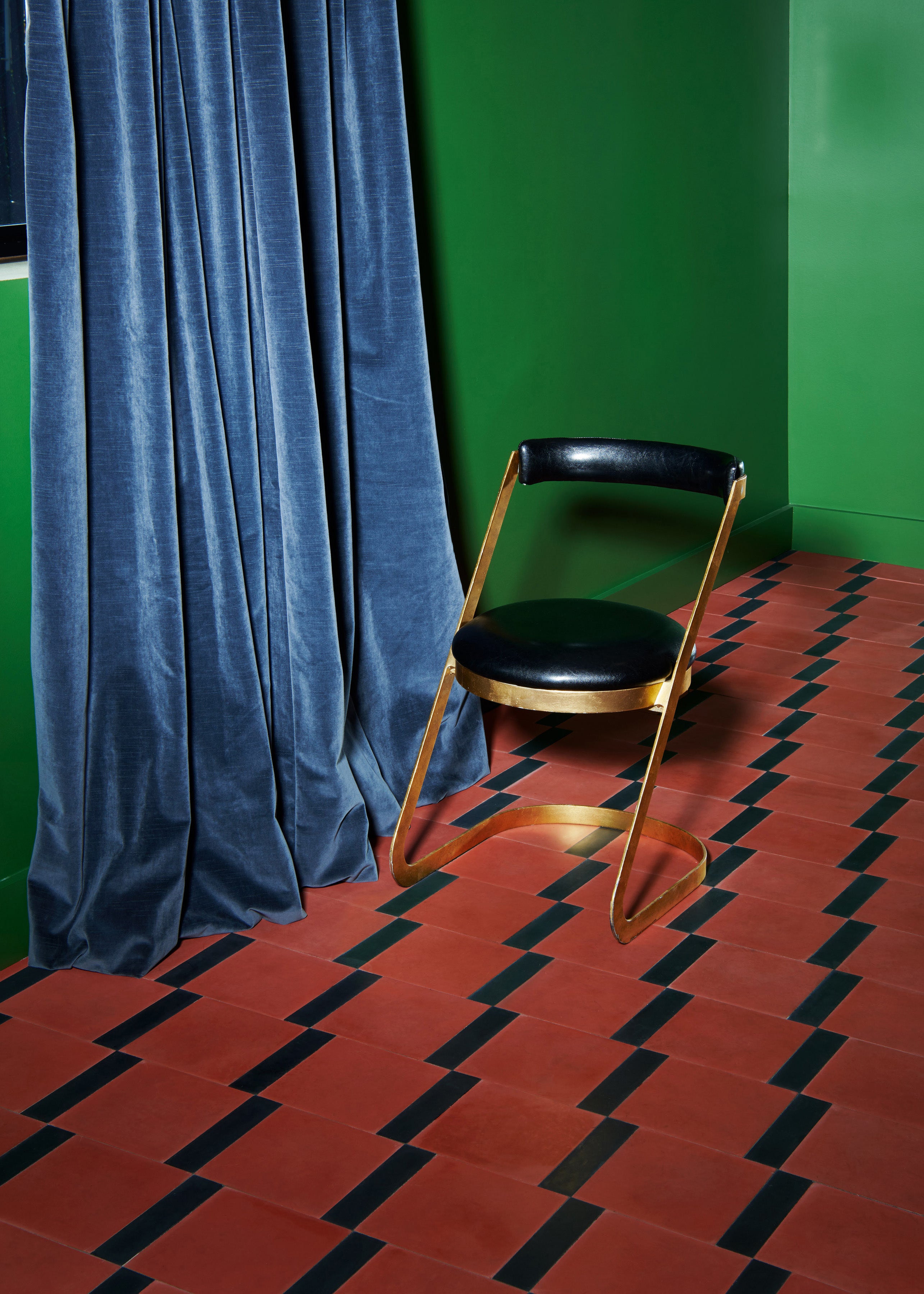 A geometric dark terracotta and black floor with a green wall, blue velvet curtain and lone gold and black vanity chair.