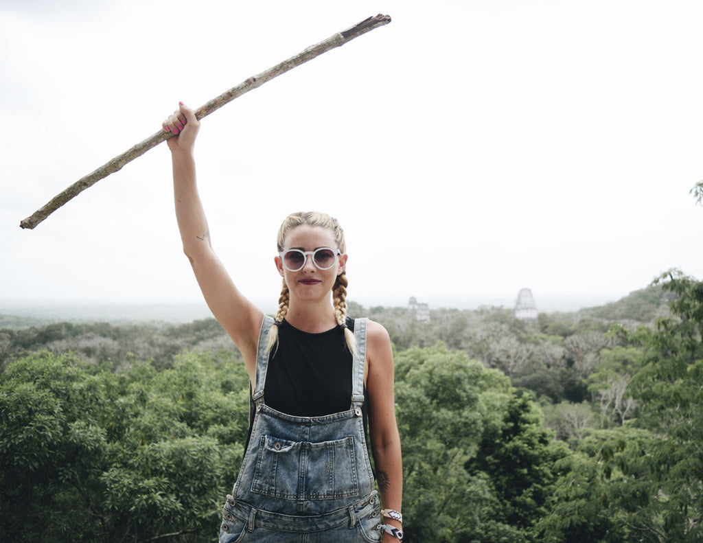 Alyssa is holding a large stick at the top of a monument and wearing overalls, a black tank top, and boxer braids
