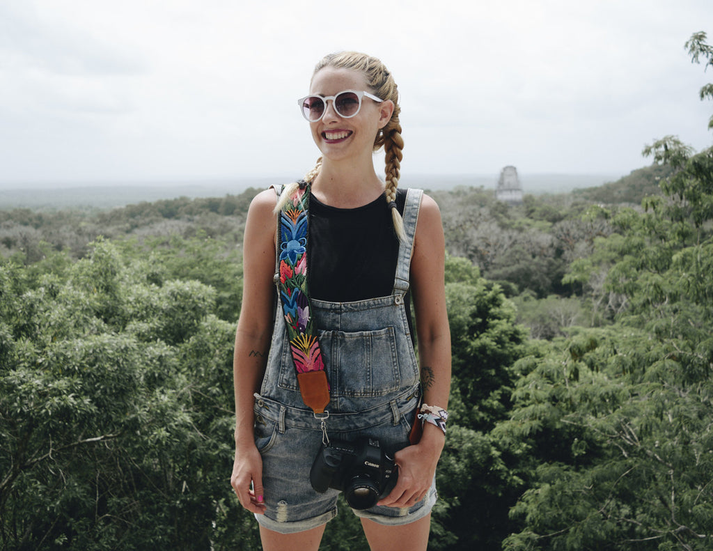 Alyssa is holding the camera with the camera strap at the top of a monument and wearing overalls, a black tank top, and boxer braids