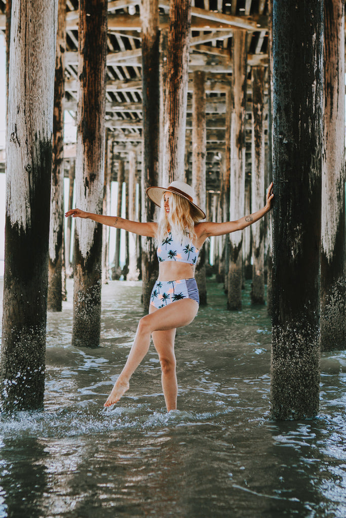 Alyssa walking under the boardwalk at the beach as she wears a palm-tree patterned bikini and a Hiptipico wide brim hat