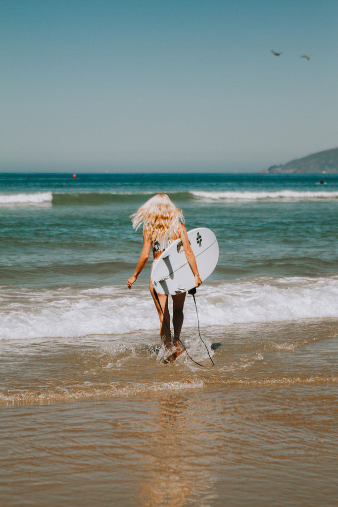 Alyssa heading towards the ocean with a surfboard in hand as she wears a palm-tree patterned bikini and a Hiptipico wide brim hat