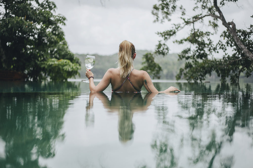 A girl with blonde hair in a swimsuit holding a glass of champagne looking out into a lake from a swimming pool. 