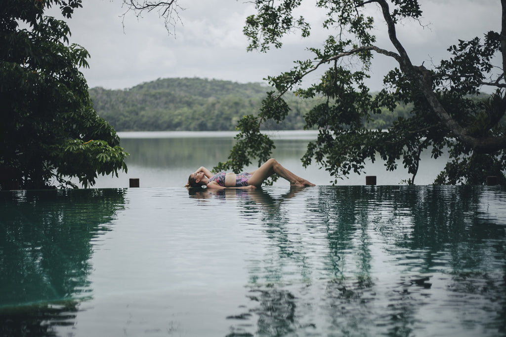 A girl with blonde hair in a swimsuit holding a glass of champagne looking out into a lake from a swimming pool. 