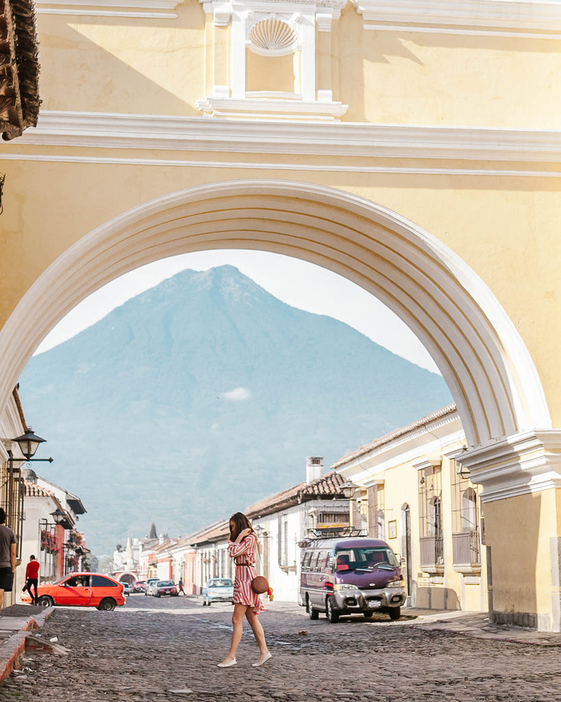 She Is Not Lost blogger Carina Otero walks under famous yellow archway in Antigua wearing Hiptipico cross body bag and red and white striped dress, Antigua travel, destinations in Guatemala, must see Guatemala, picturesque locations in Guatemala, ethical travel bloggers