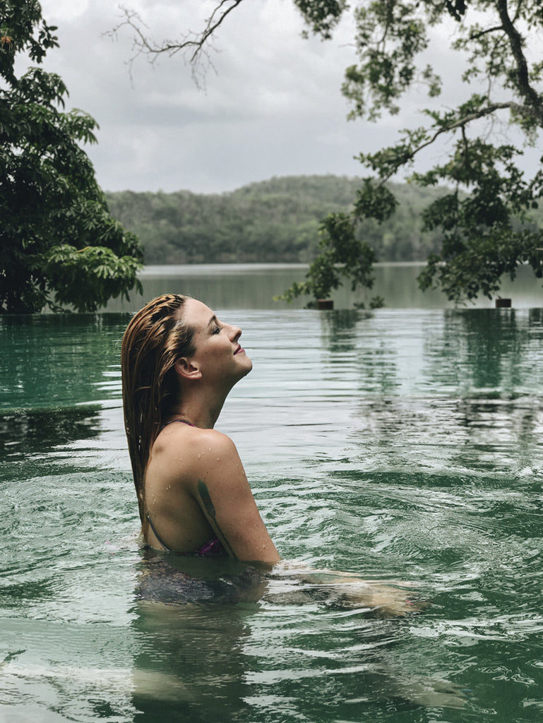 A girl with blonde hair in a swimsuit holding a glass of champagne looking out into a lake from a swimming pool. 