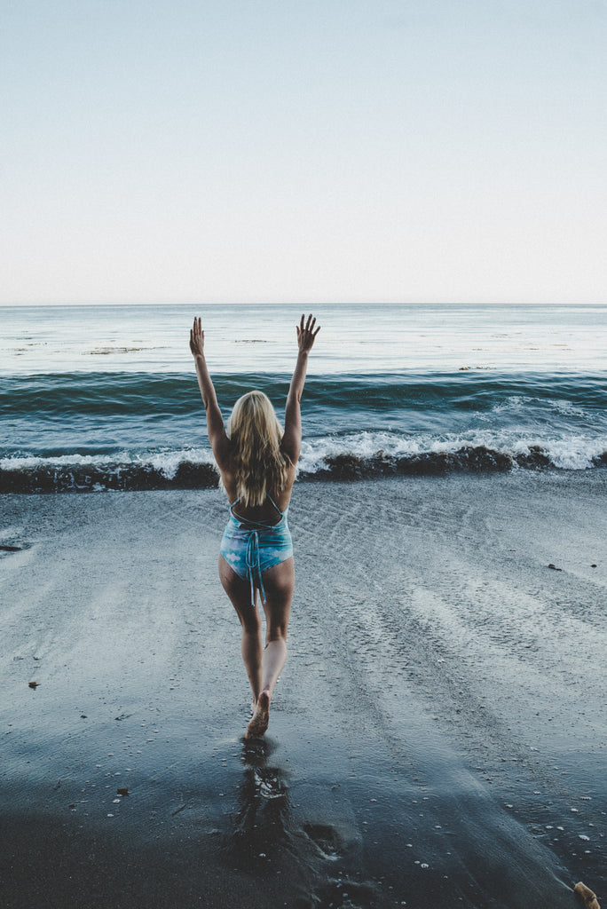 Alyssa facing the ocean with arms tossed up in the air wearing a blue and white tie-dye one piece swimsuit.