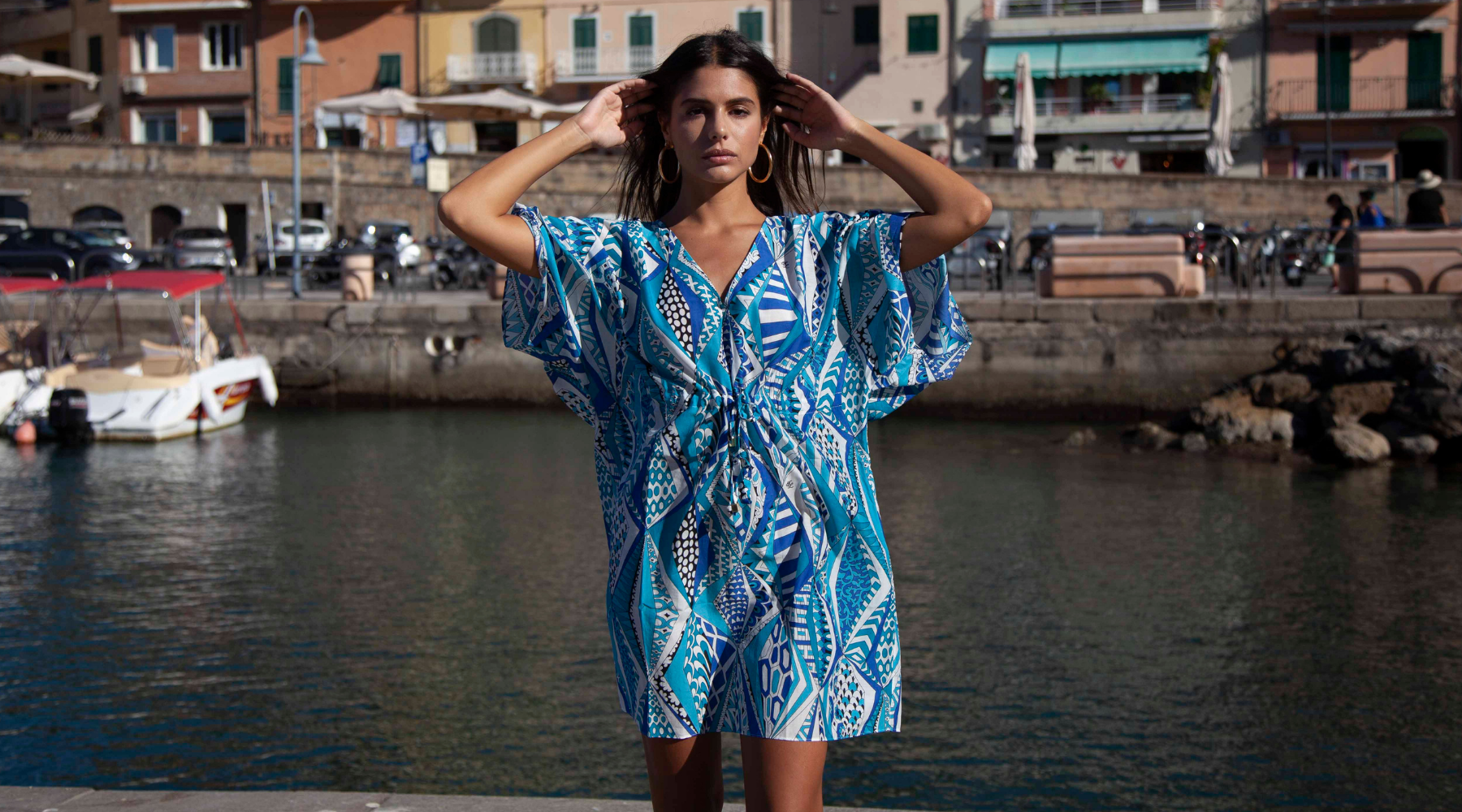 Model poses at waterfront with boats and buildings in the background, wearing the Nieves Lavi Riva Kaftan in Capri Print, a vivd pattern of blues, whites, and black