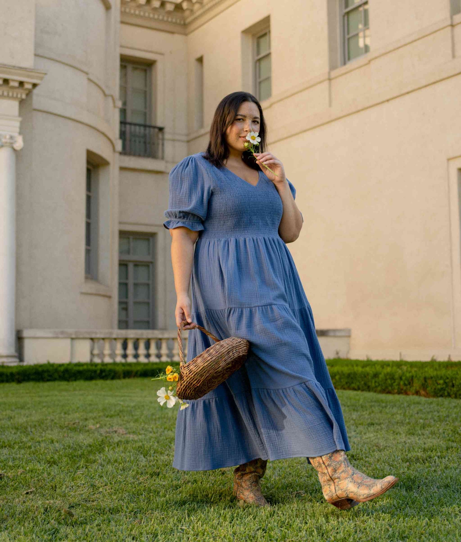 Model outside wearing blue dress from Christy Dawn, holding a basket and sniffing a flower.